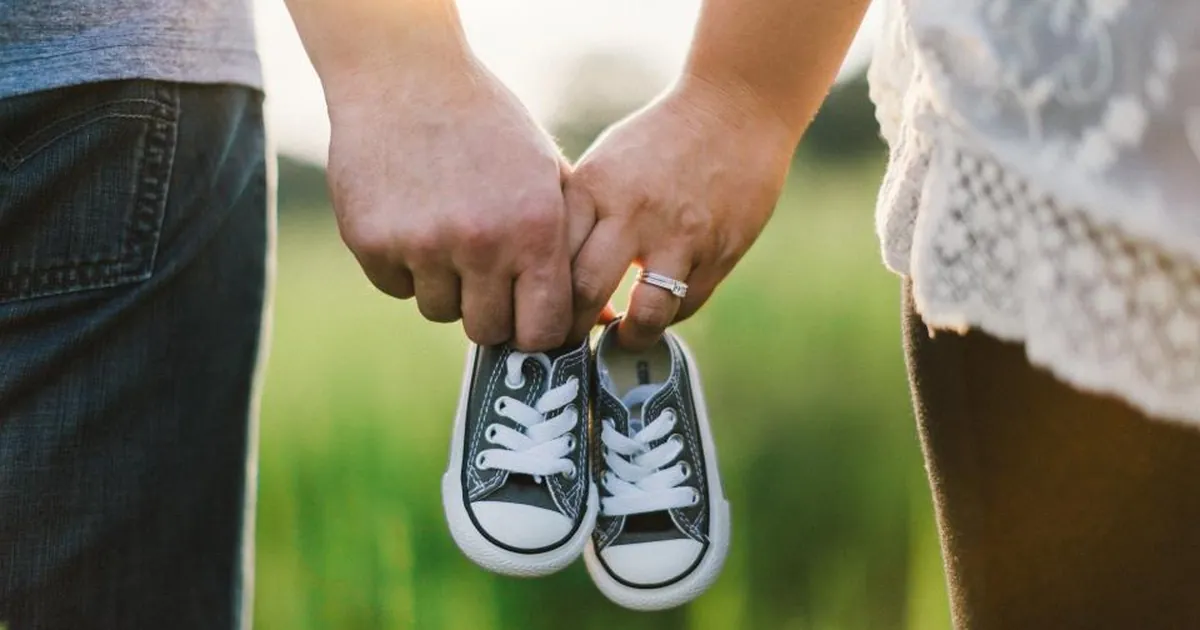 Two parents hold a pair of small baby shoes between them in a green field.