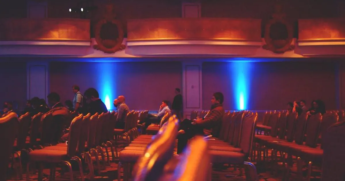 Guests take their seats in a conference hall with red chairs and blue stage lights.