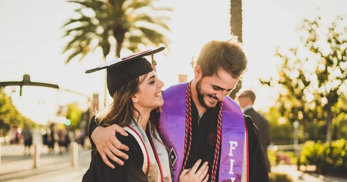 Two graduates in caps and gowns hug and laugh on a sunny street.