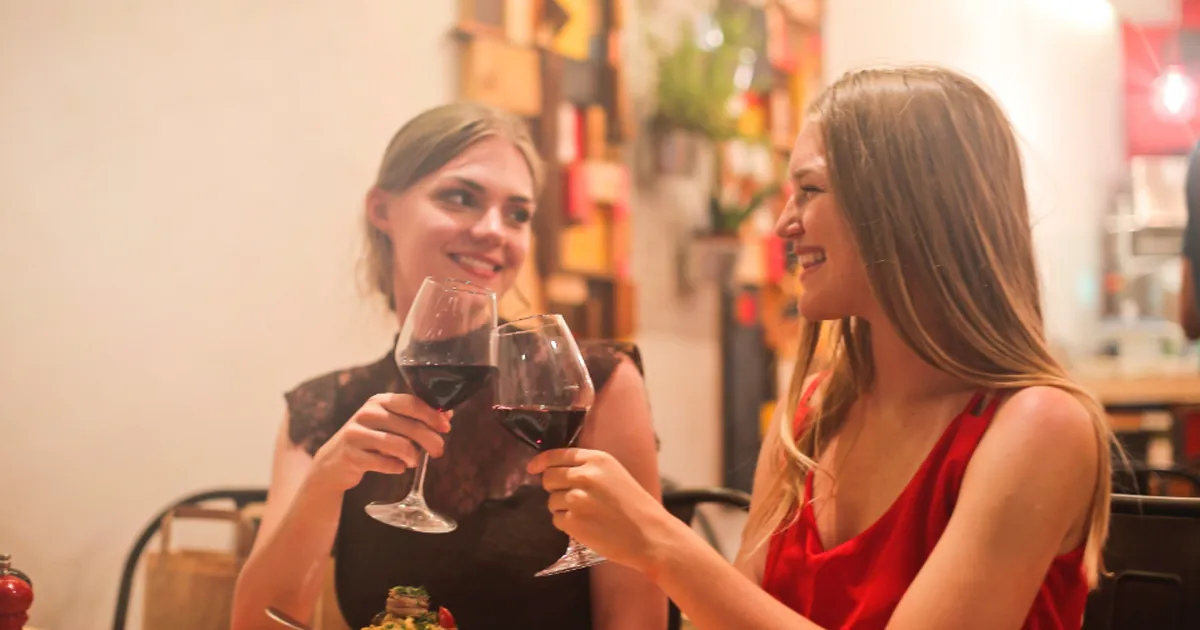 Two friends smile and touch their glasses of red wine at a restaurant table.